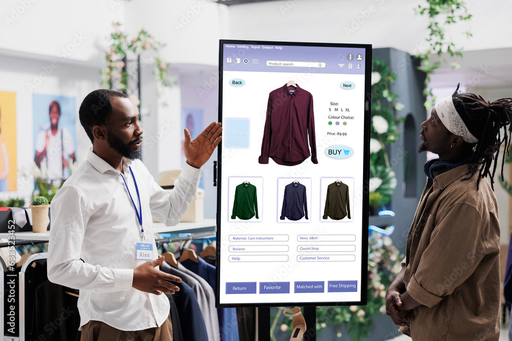 Male assistant showing clothes on interactive kiosk board to young trendy customer in clothing store at mall. Retail employee helping client to choose modern fashion items, self ordering.